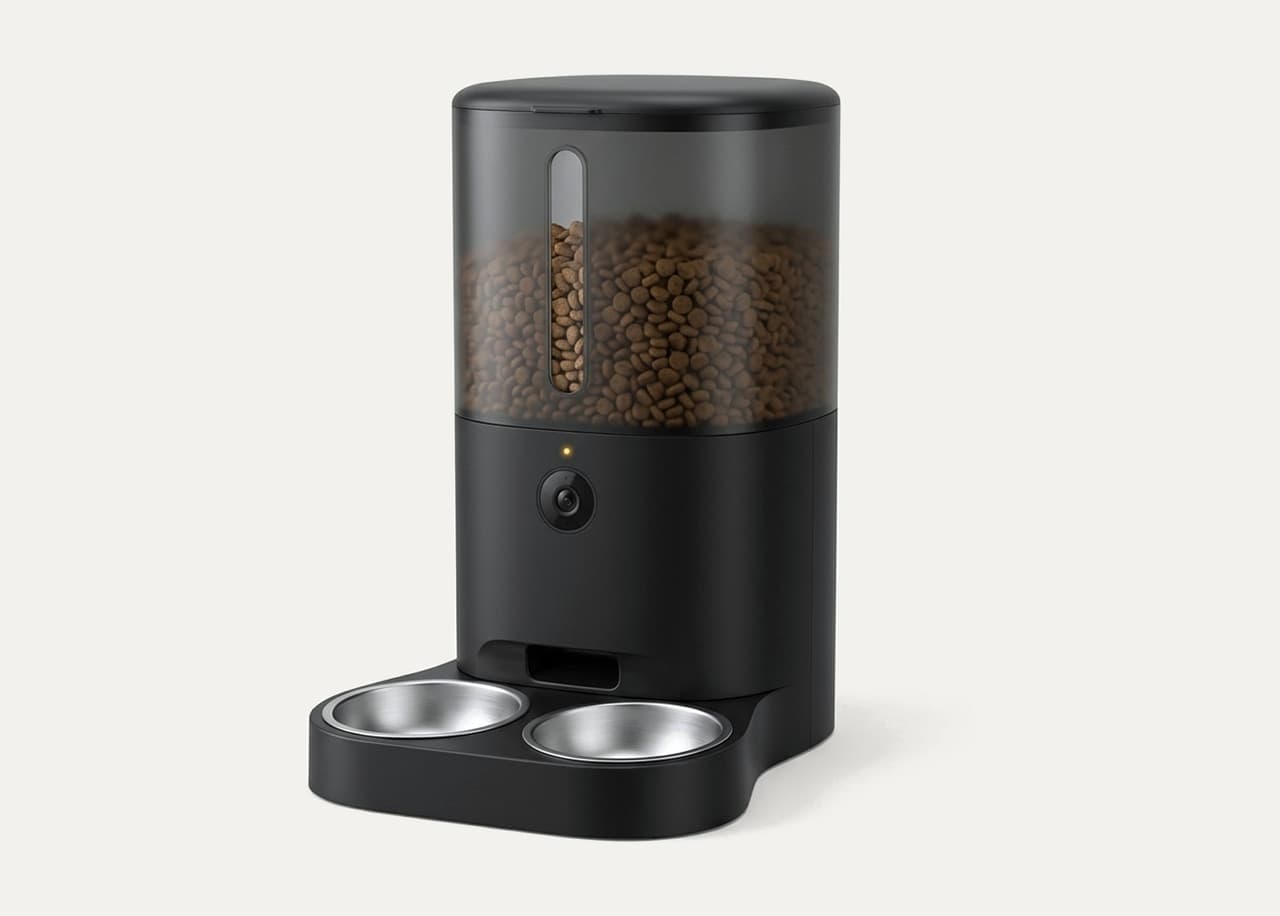 Distributeur de croquettes noir avec caméra intégrée et double gamelle inox, croquettes visibles dans le réservoir, sur fond ivoire
